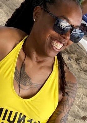 Sakhara is basking in the Virgin Island sun at the beach as she smiles for a photo. She is wearing a yellow one piece swimsuit with black lettering. She is covered in tattoos, wears clip-on sunglasses over her prescription lenses and studded earrings.