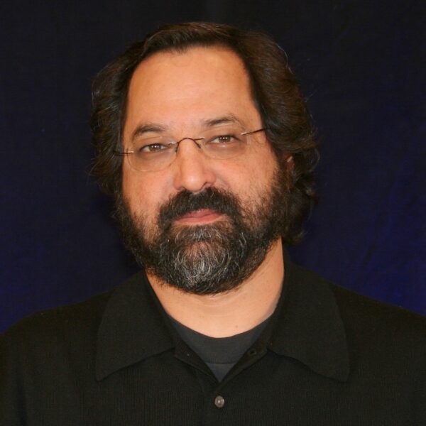 Dr. Michael Perlis, a white man with dark flowing hair and a beard wearing unframed glasses and a black polo shirt.