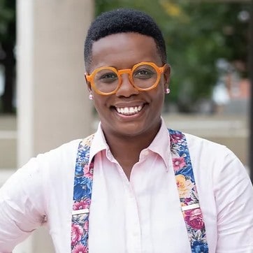 Dr. Kali Cyrus, a black person wearing bright orange framed glasses, a white button down shirt, and floral printed suspenders.