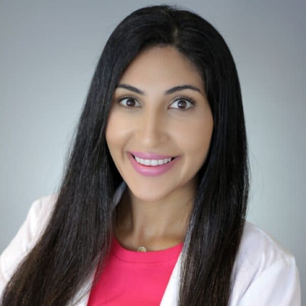 Dr. Nora Ghodousi-Zaghi, a woman with long, dark hair and olive-toned skin wearing a pink top and white coat.