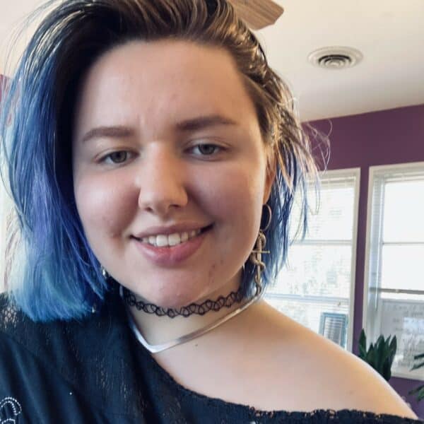 Rachel Aubry, a person with chin-length dyed blue hair, wearing an off-the-shoulder top and jewelry.
