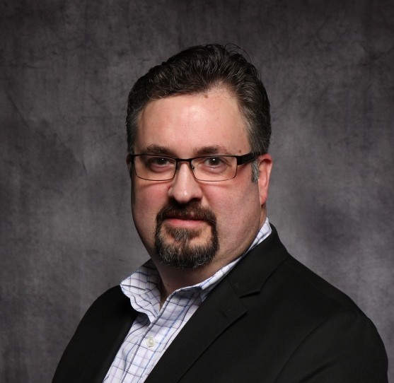 Ed Pardo, a man with dark hair and a goatee wearing glasses, a button-down shirt. and black jacket posing for a professional headshot.